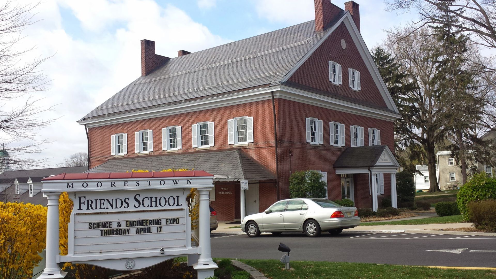 Friends School sign in front of a red brick school building with a gray roof and parked cars.
