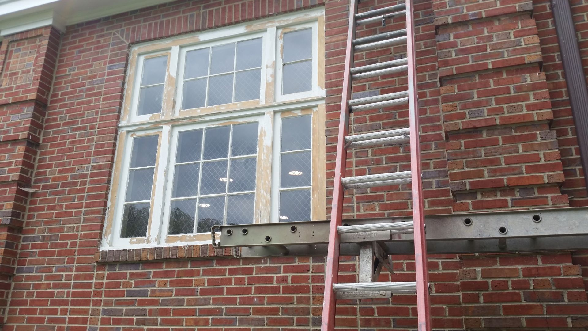 White-framed window on a red brick wall with metal supports beneath it and a ladder or pipe on the right.