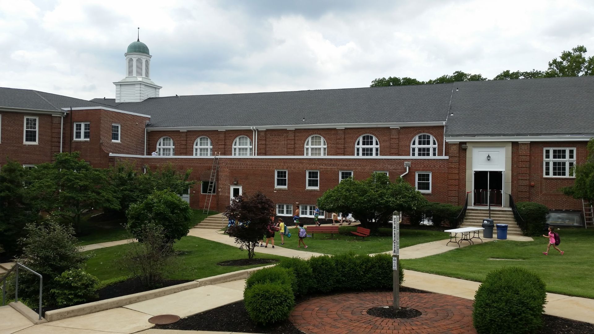 Brick office building with white cupola, landscaped courtyard, and walkways under a cloudy sky