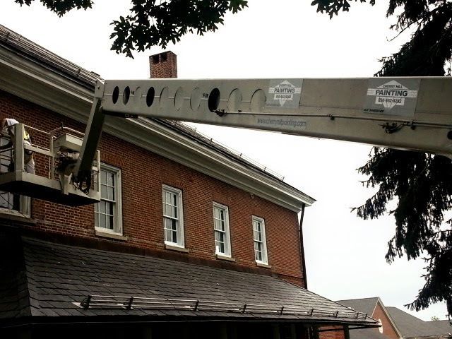 Bucket truck boom beside a red brick building with white-trimmed windows and overhanging tree branches