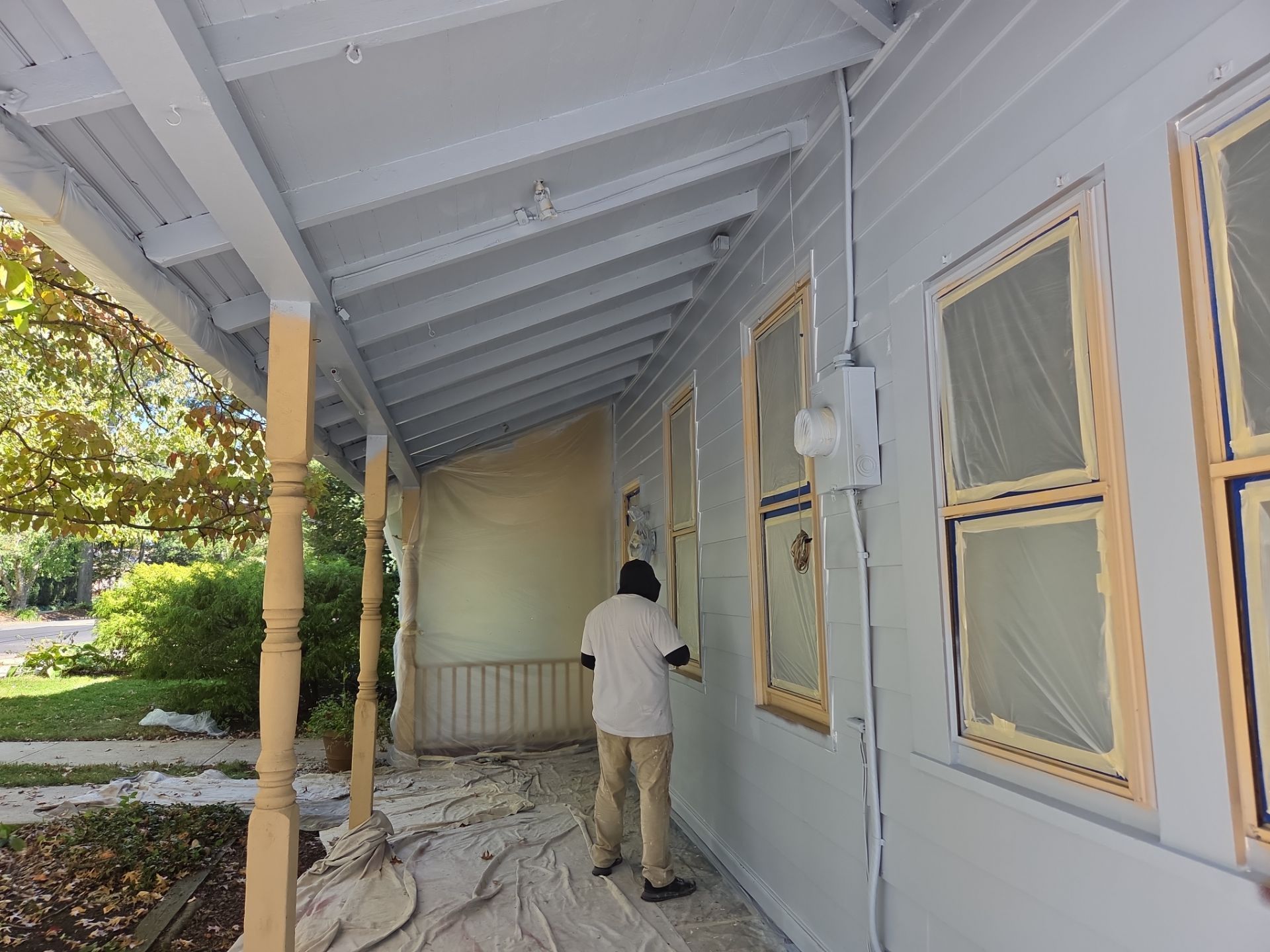 Person painting a porch wall under a covered roof beside windows and yellow columns