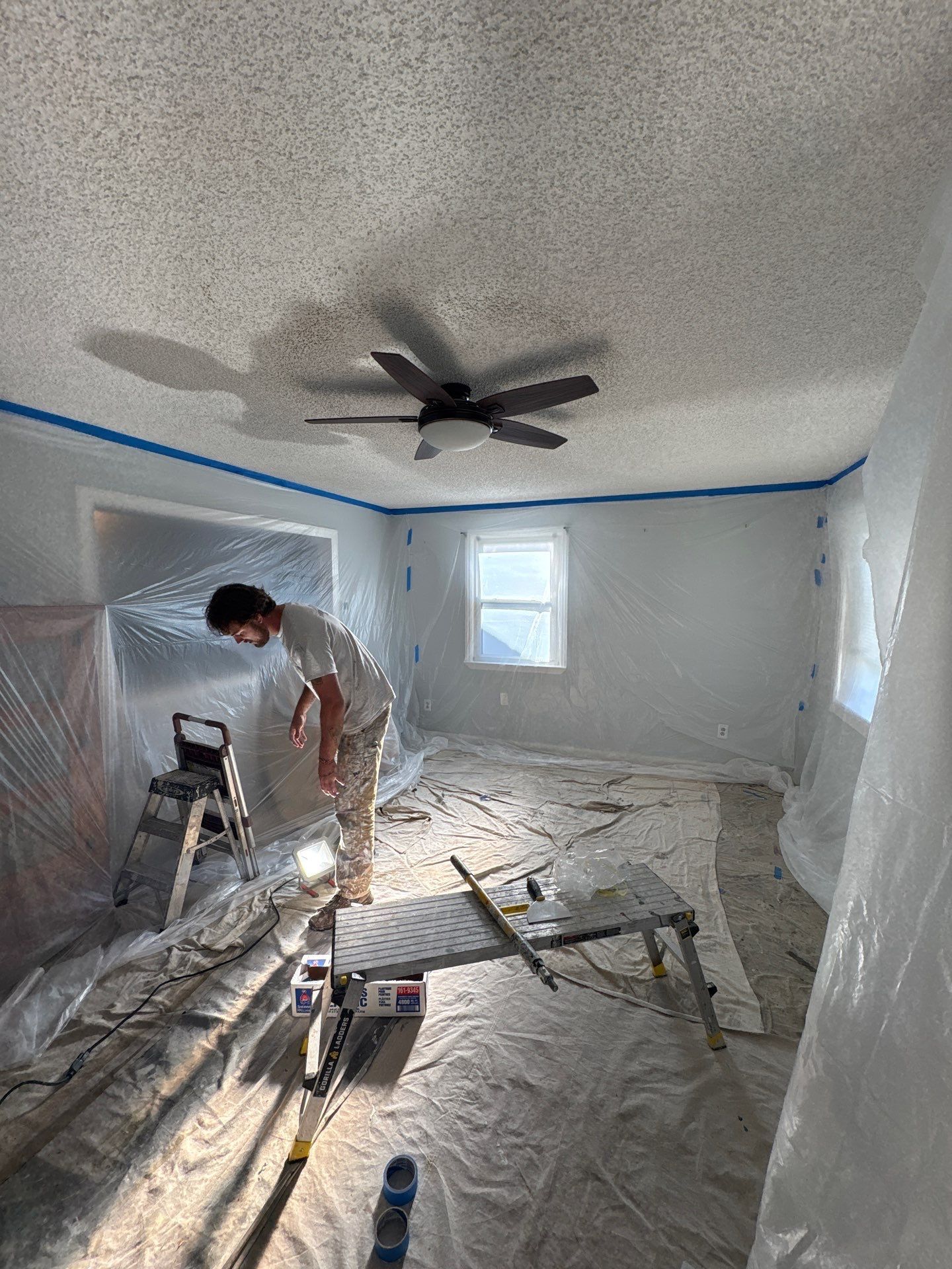 Person painting a room wrapped in plastic sheeting, with a ceiling fan and tools on the floor.