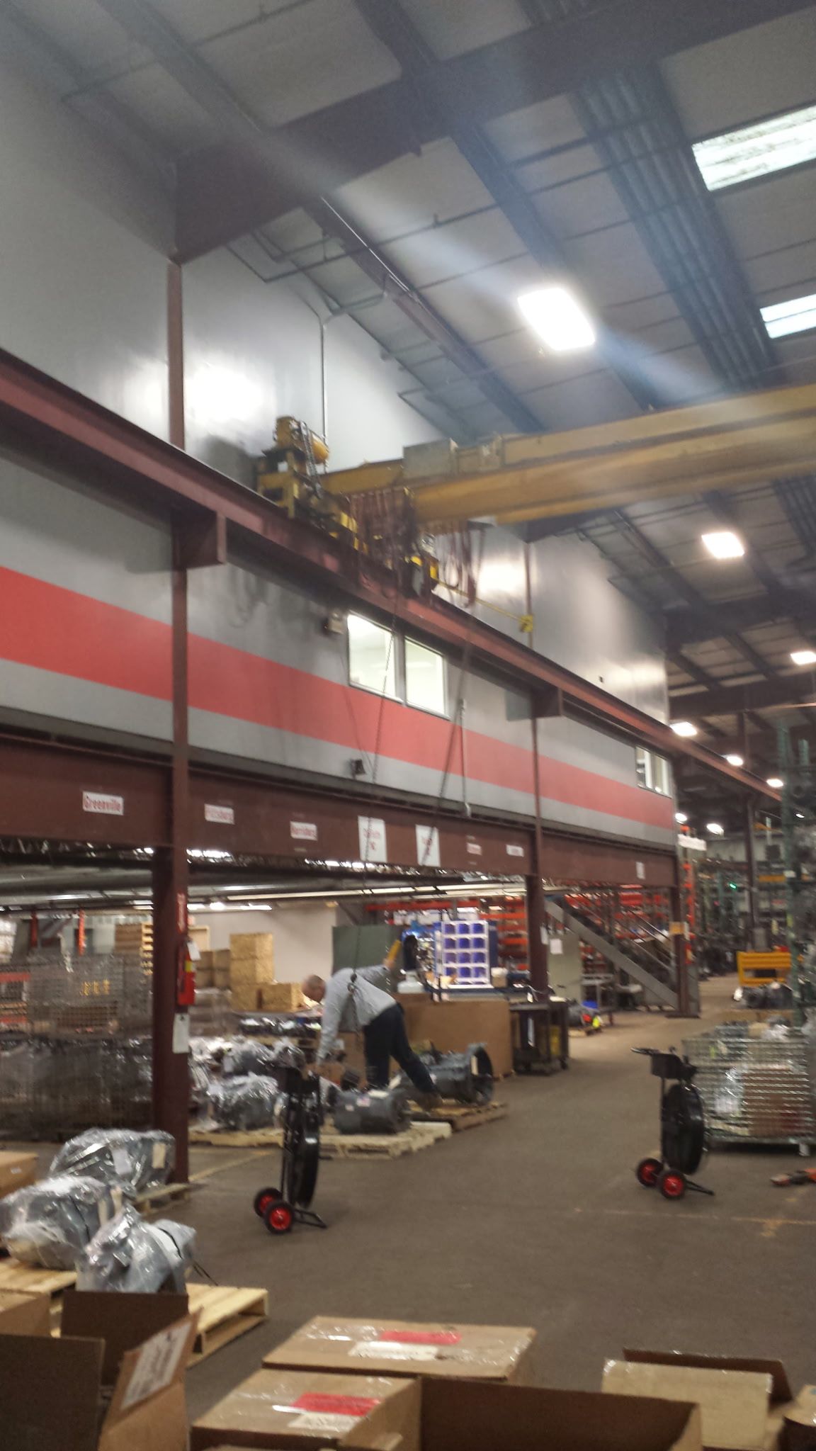 Large industrial warehouse with overhead crane, stacked boxes, and workers among pallets and equipment.