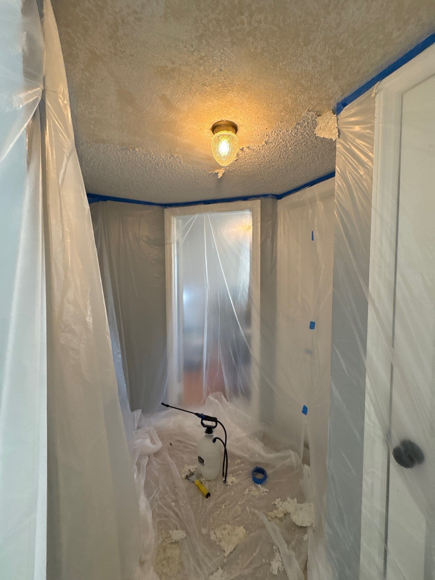 Plastic-covered hallway under renovation with drop cloths, blue painter’s tape, and a ceiling light.