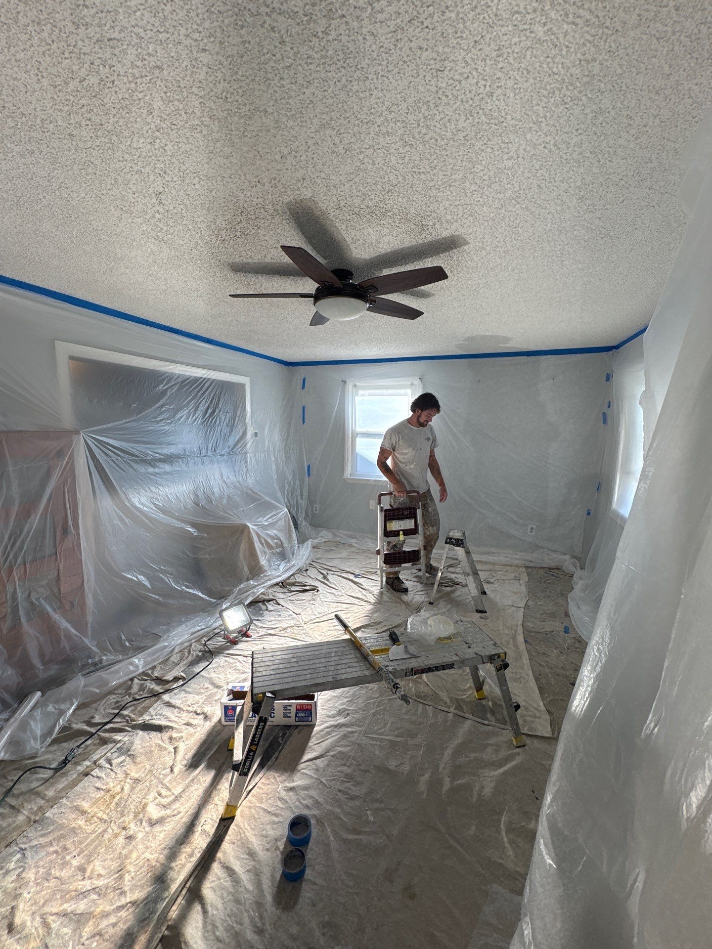 Worker painting a room under renovation, with plastic-covered walls, ceiling fan, and tools on the floor.