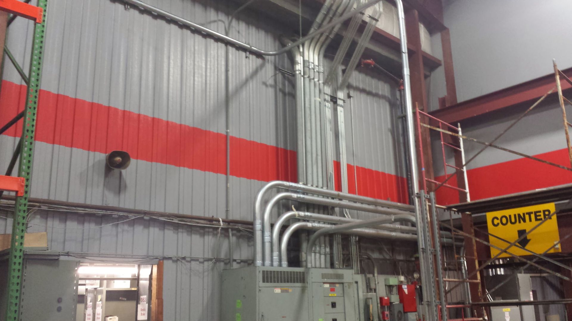 Industrial interior with red and gray walls, silver pipes, and a yellow “COURTESY” sign beside scaffolding.