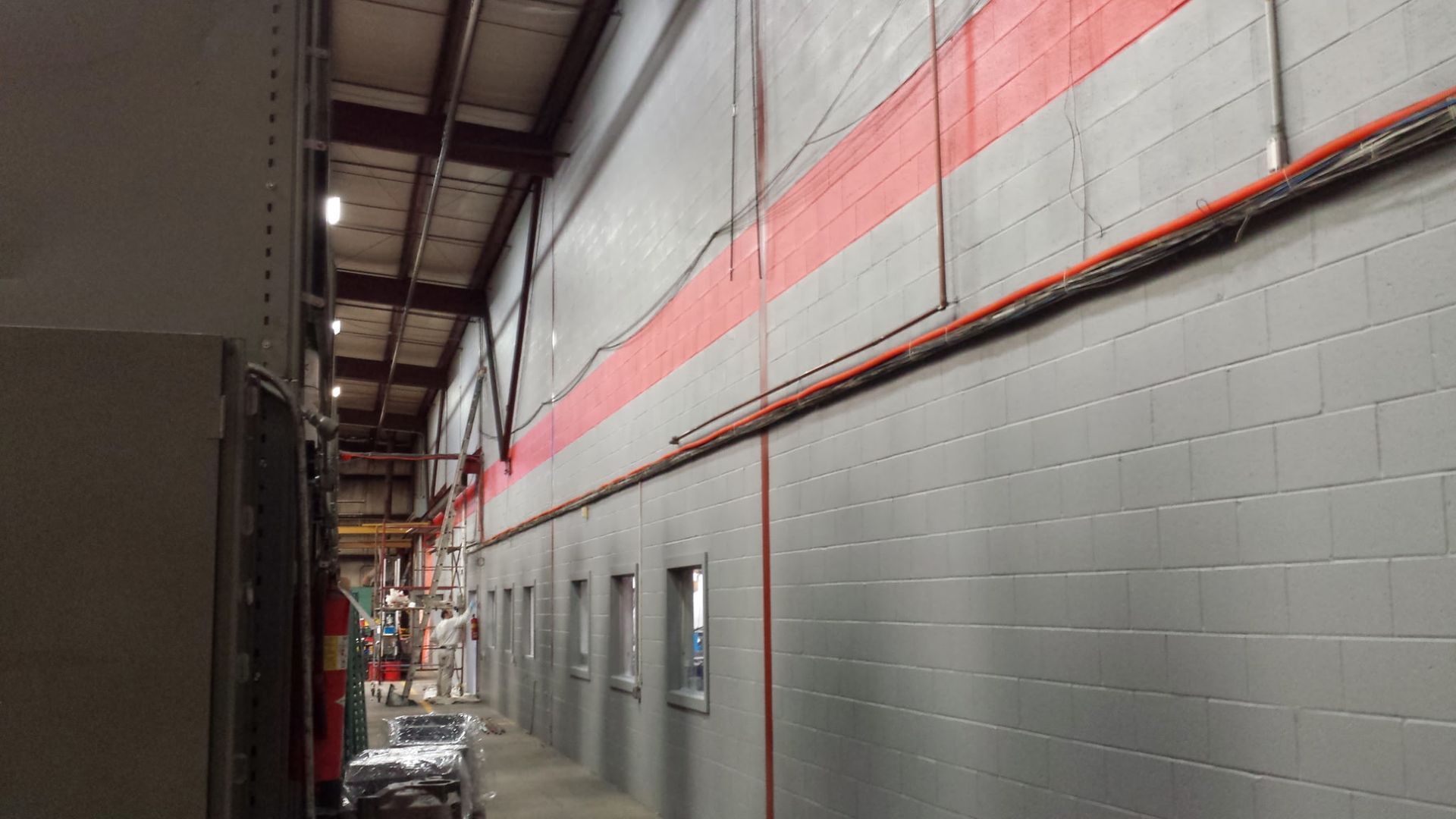 Narrow industrial hallway with gray cinderblock walls, red trim, and overhead piping in a warehouse-like building