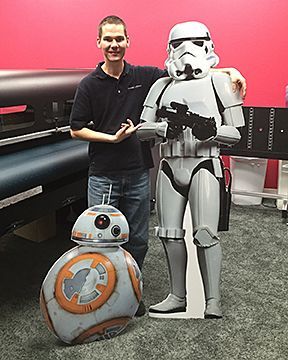 A man is standing next to a cardboard cutout of a storm trooper and a bb-8 robot.