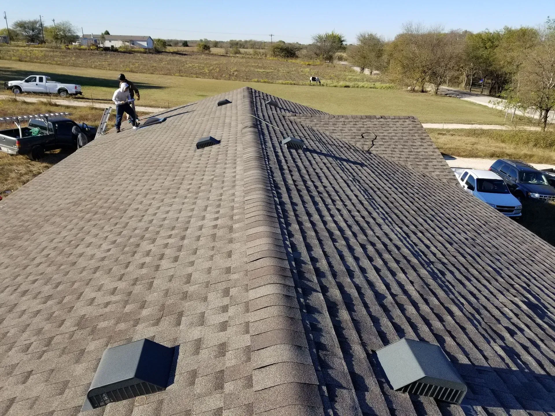 A roof with gray shingles. A worker is standing on the roof, other vehicles and fields in the background.