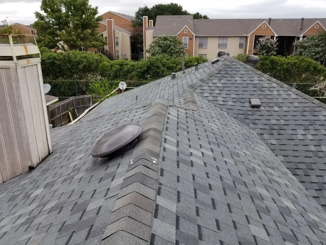Gray shingle roof with a dark, oval vent, overlooking buildings and trees.