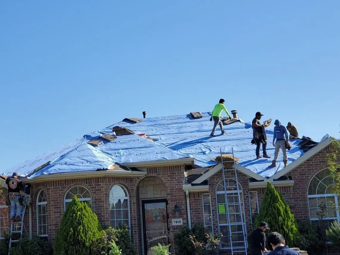 Roofers working on a house roof. Blue tarp covers parts of the damaged roof on a sunny day.