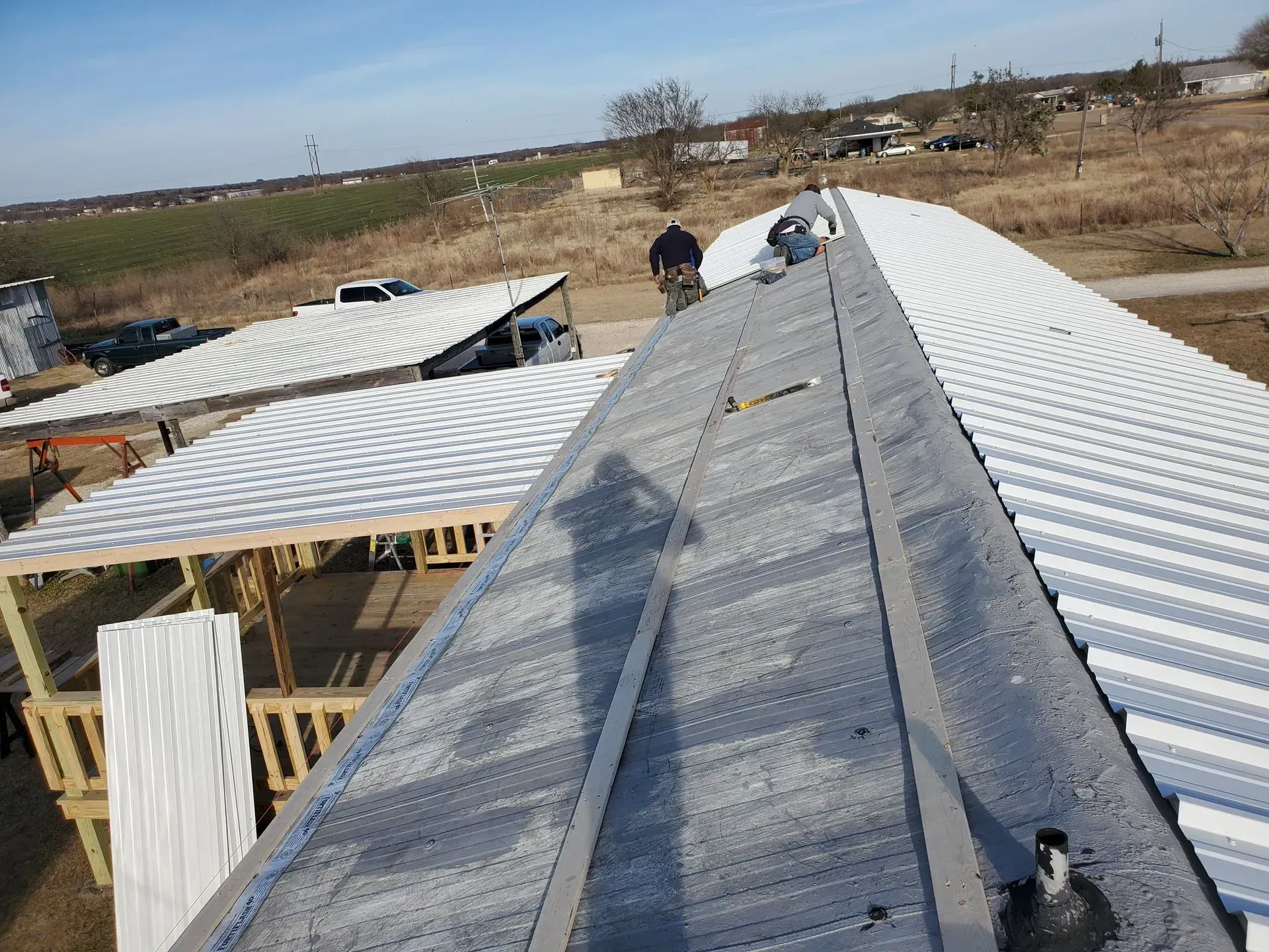 Workers installing a metal roof on a building, another building visible. Outdoors.