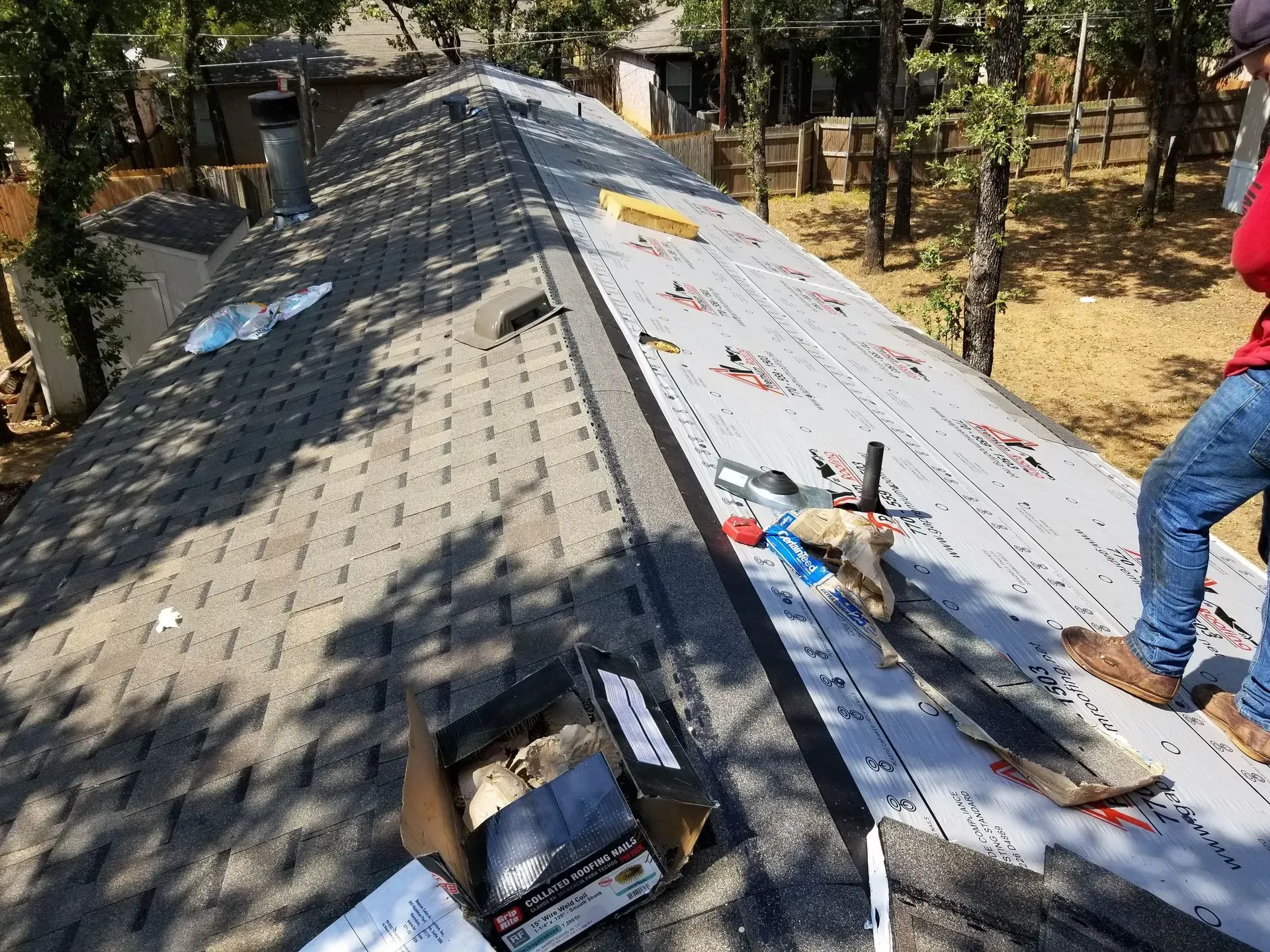 Roofer on a roof installing shingles. Gray shingles, underlayment, and tools are visible. Sunny outdoor setting.