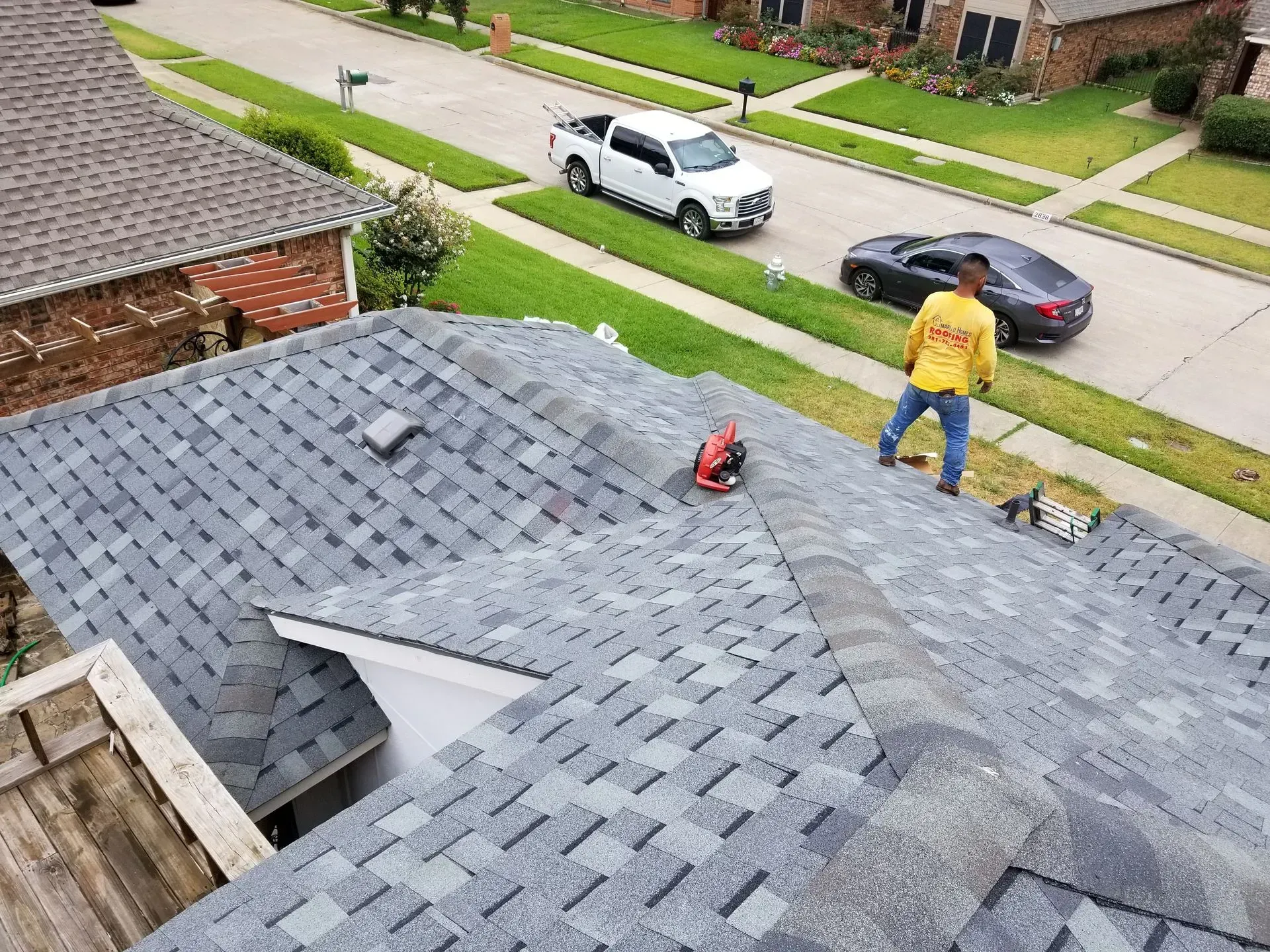 Roofer on gray shingle roof, cutting material. Trucks and houses in background, overcast day.