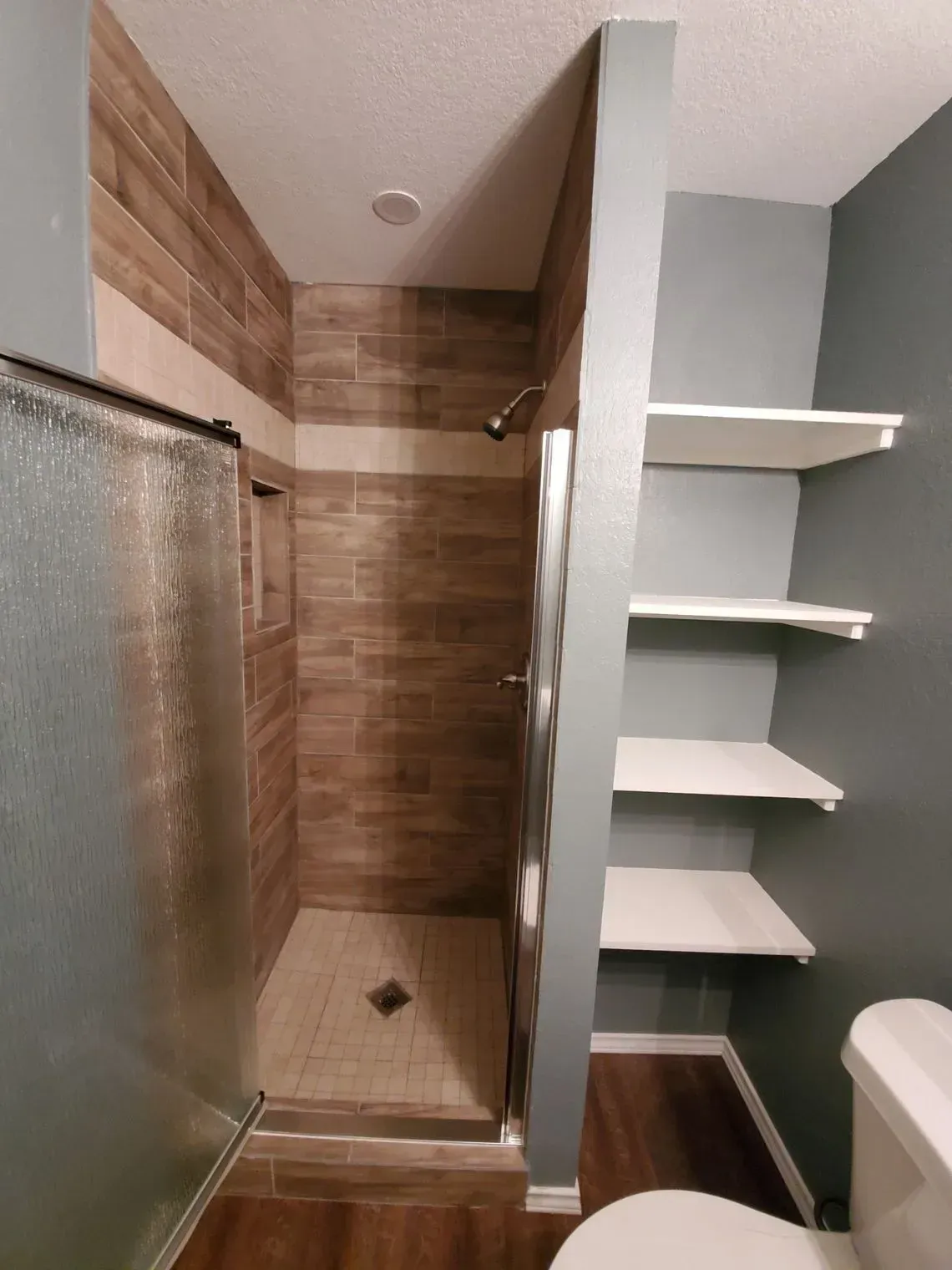 Bathroom with a tiled shower, glass door, and built-in shelves on the wall, next to a toilet.