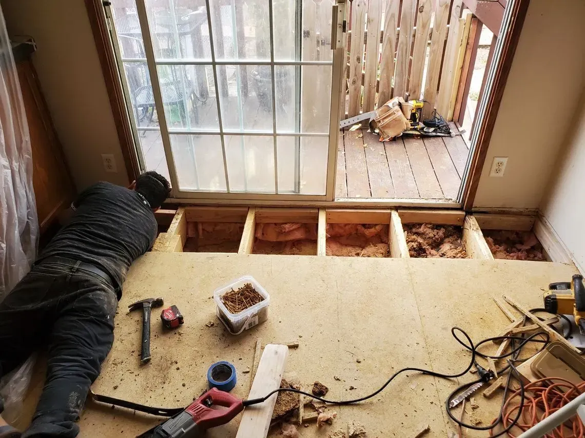 A person removing flooring near a sliding glass door and balcony. Tools and debris are scattered on the floor.