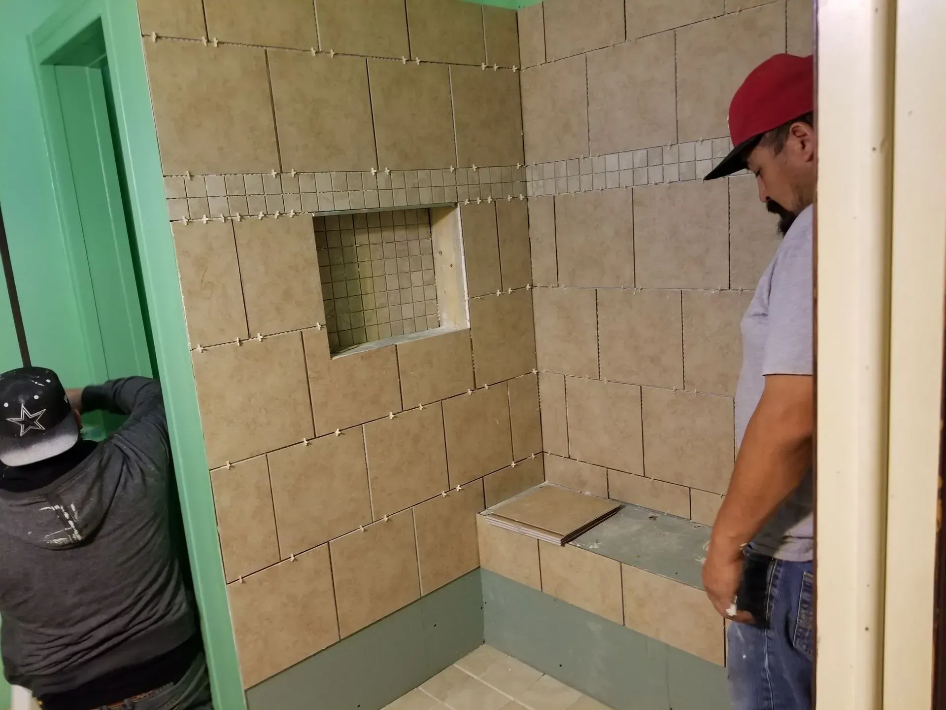 Two people tiling a shower; beige tiles, green walls, red cap, doorway.