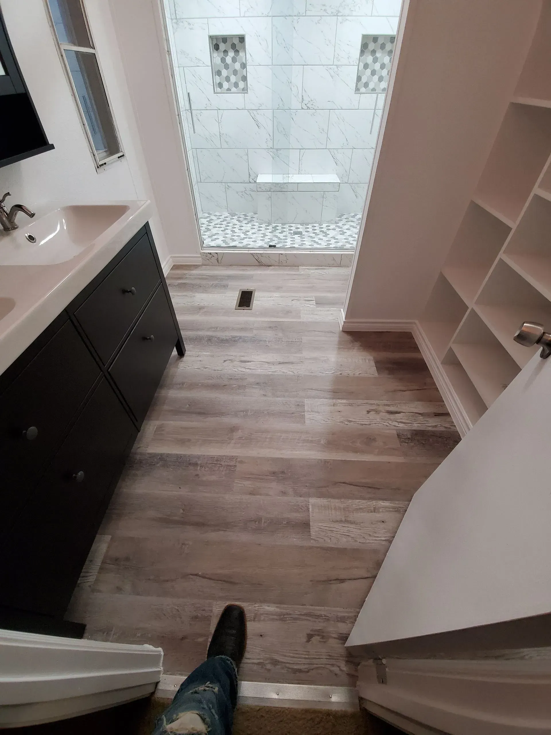 Bathroom with wood-look flooring, vanity, shower, and built-in shelving. A person stands in the doorway.