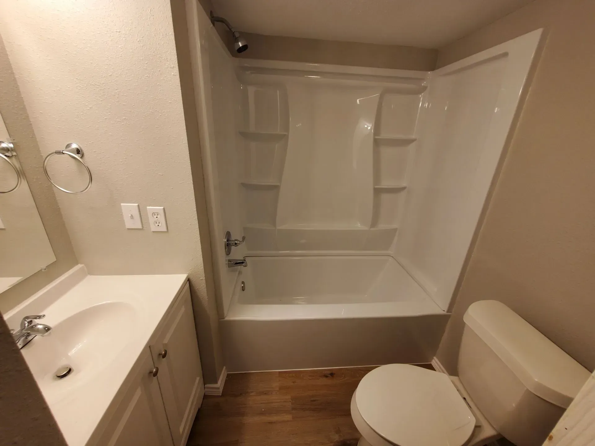 Bathroom with white tub/shower combo, toilet, sink, and cabinets. Brown wood-look floor, tan walls.