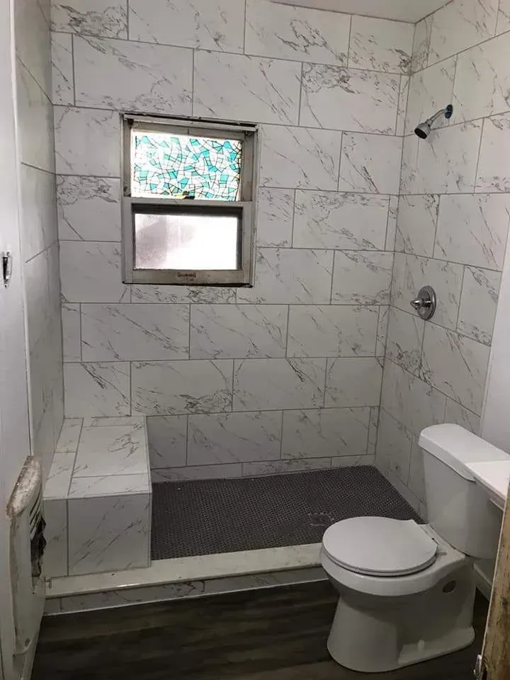 Bathroom with white marble-like tile walls, a toilet, a showerhead, and a window above a built-in bench.