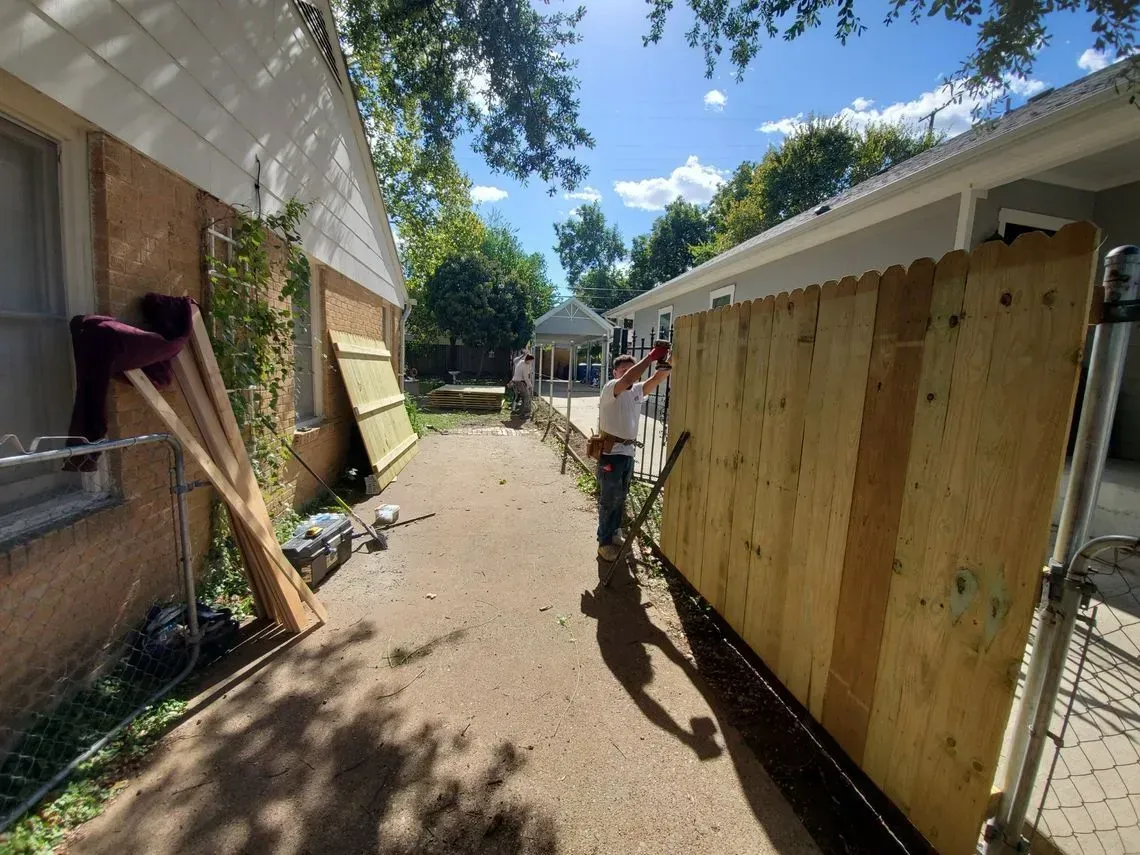 Person builds wooden fence in narrow outdoor alleyway between buildings on sunny day.