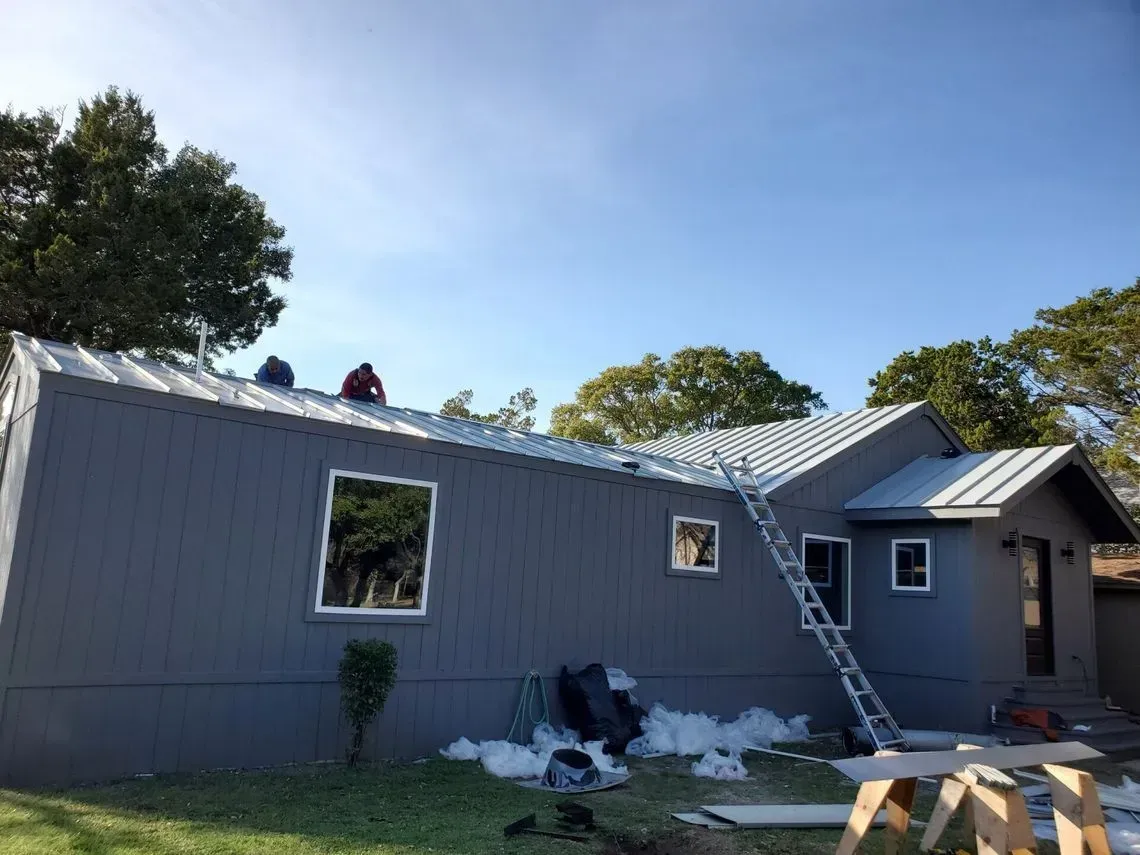 Workers installing a metal roof on a gray house under a blue sky. A ladder is leaning against the roof.