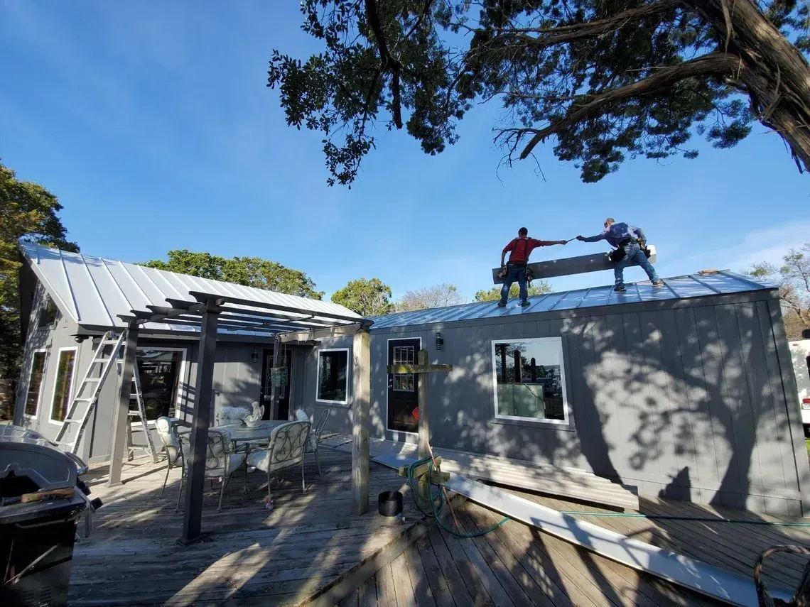 Two people on a gray roof, working. A house with a gray exterior sits on a wooden deck under a blue sky.