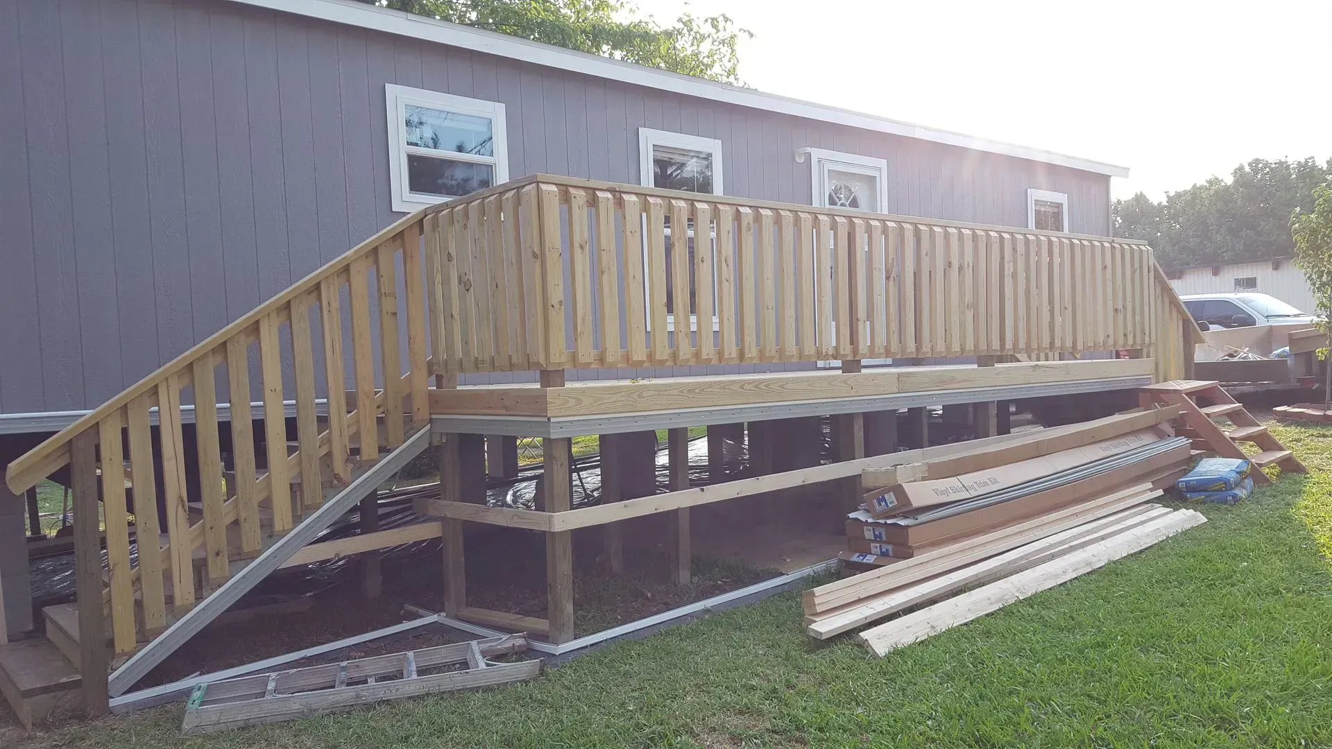 Wooden deck with railing and stairs attached to a gray house, set on cinder blocks over grass.