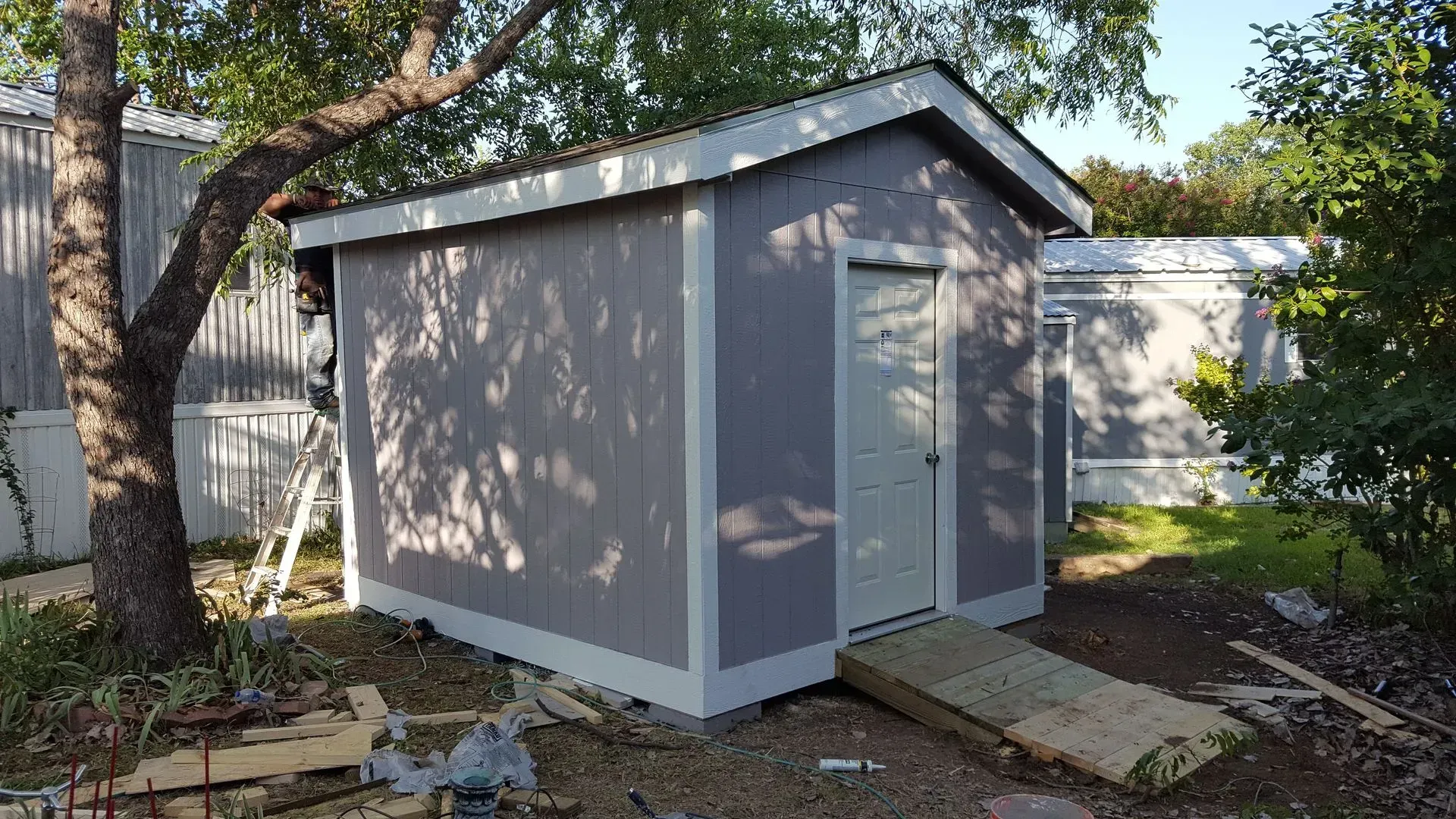 A gray shed with a white door and ramp, set outside with a tree nearby.