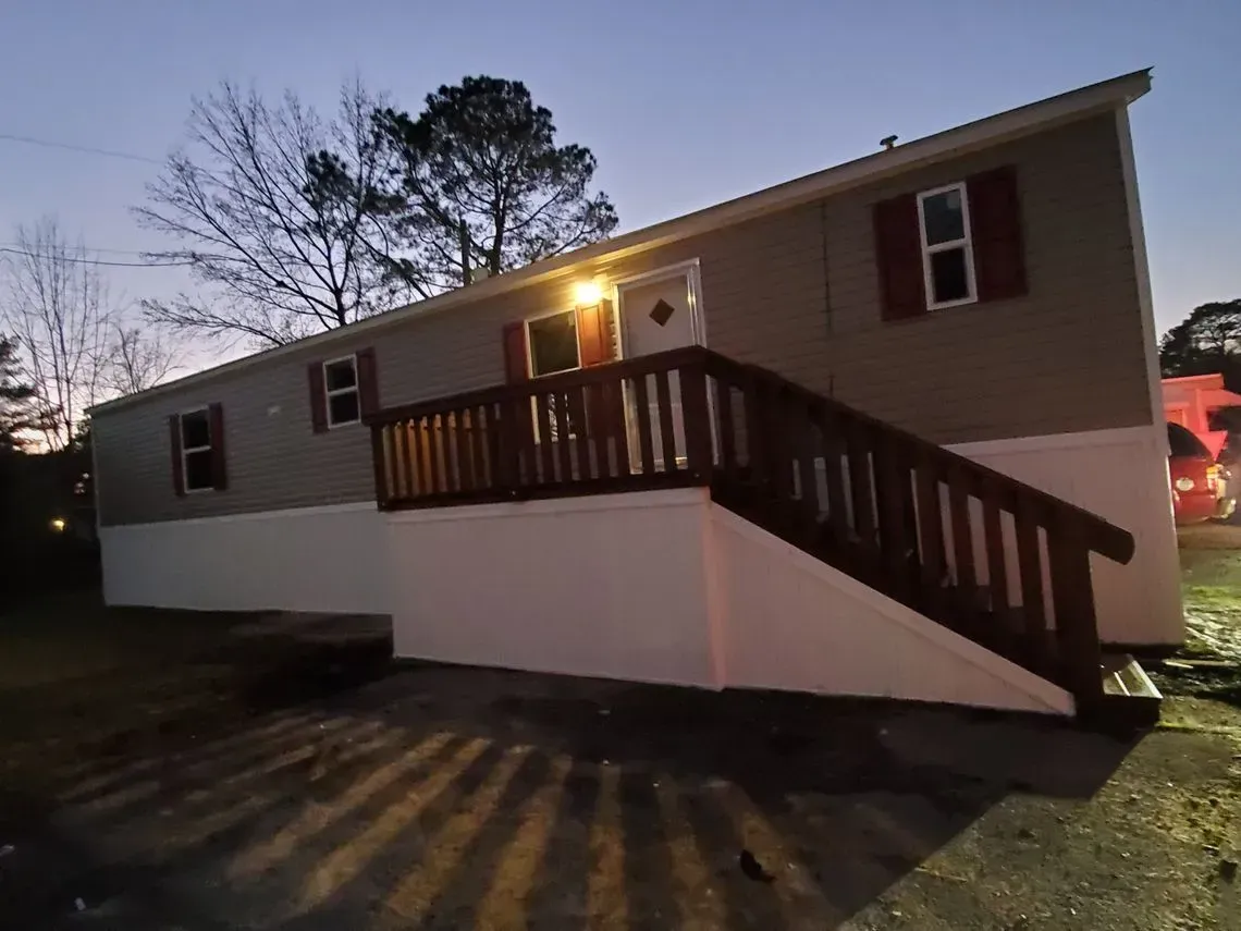 A light-colored, single-story house with a brown railing, red shutters, and white trim at dusk.