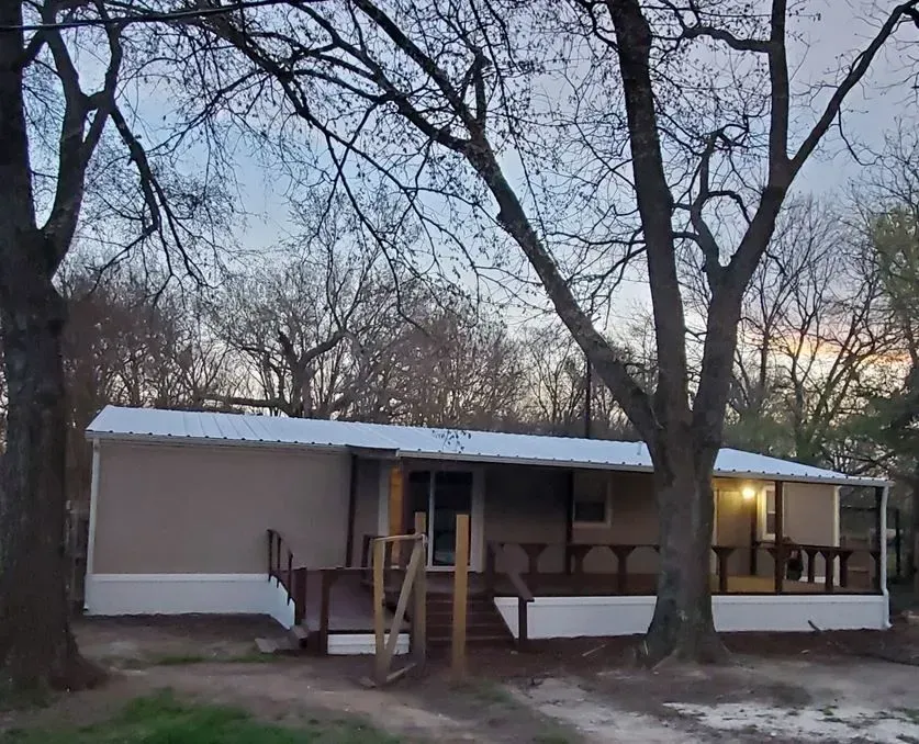 Tan single-story house with a porch and deck, surrounded by trees.
