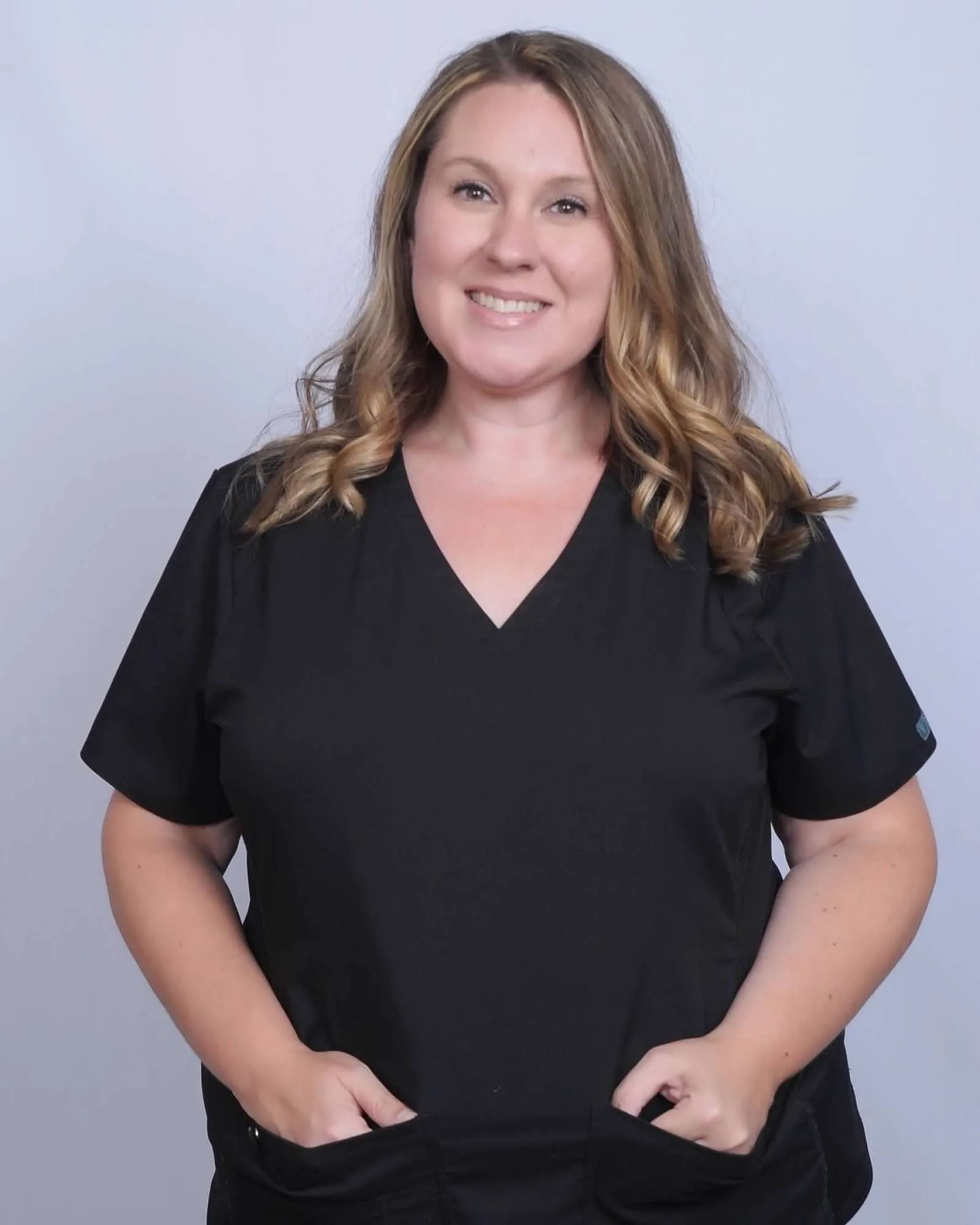 Woman with light brown hair, wearing black scrubs, smiling, hands in pockets.