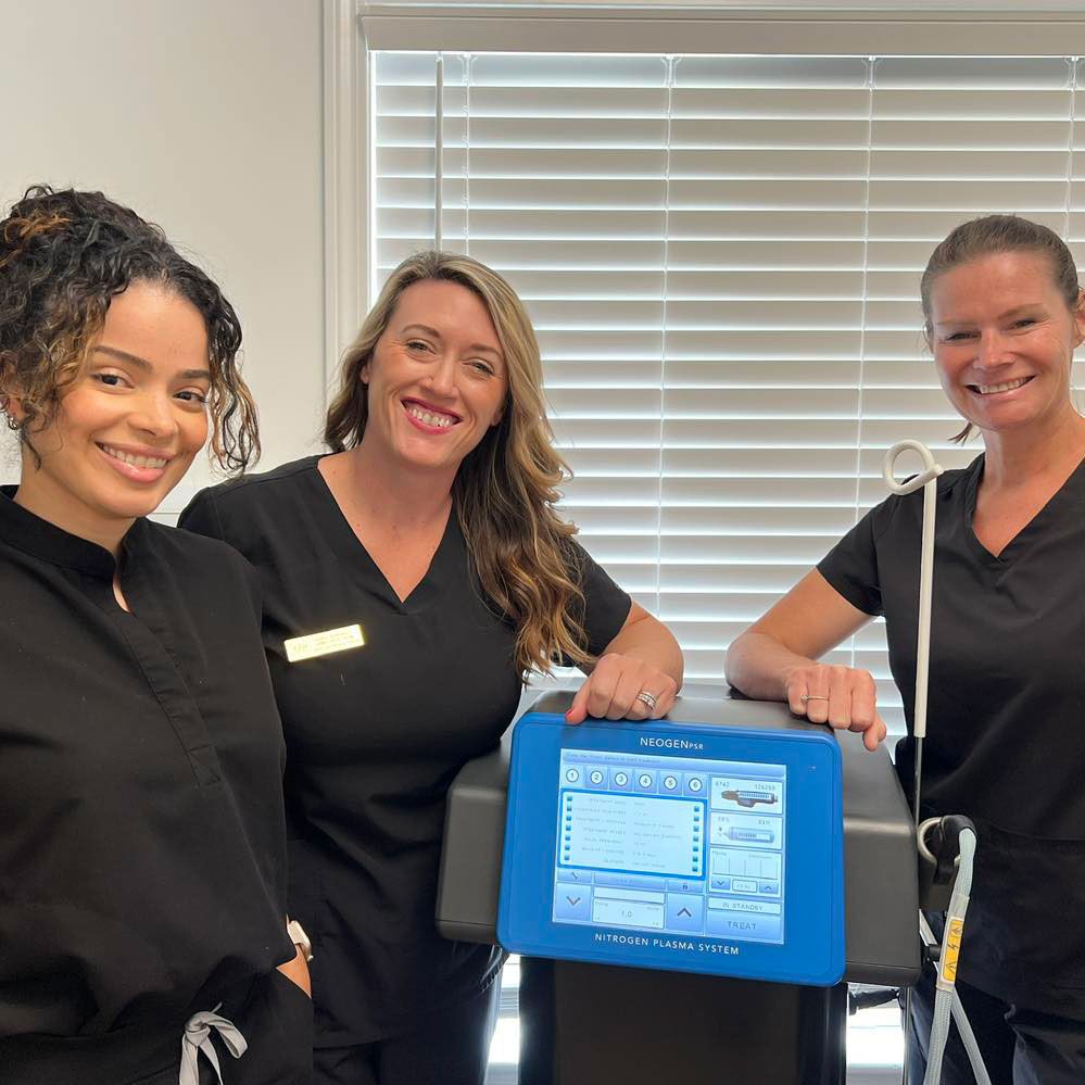 Three women in black scrubs smile next to a blue medical device in a brightly lit office.
