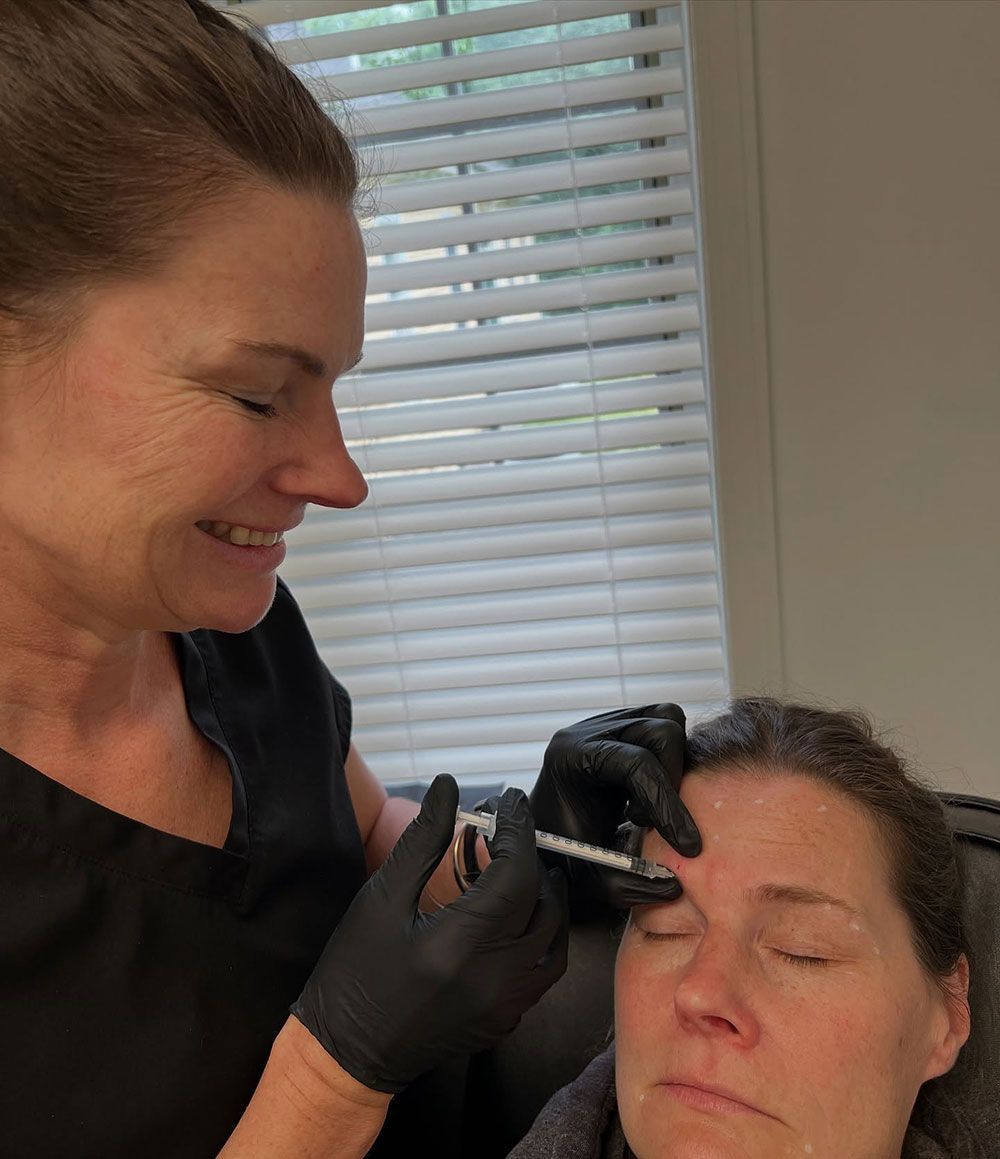 Person administering an injection to a patient's forehead; both are indoors near a window.