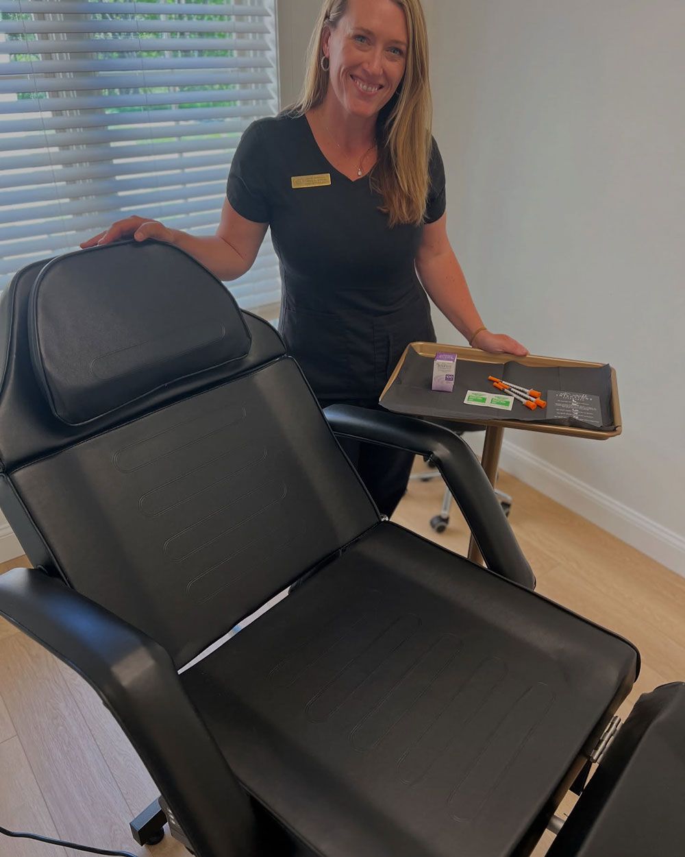 Woman smiles, holding tray of supplies beside black treatment chair in a room with blinds.
