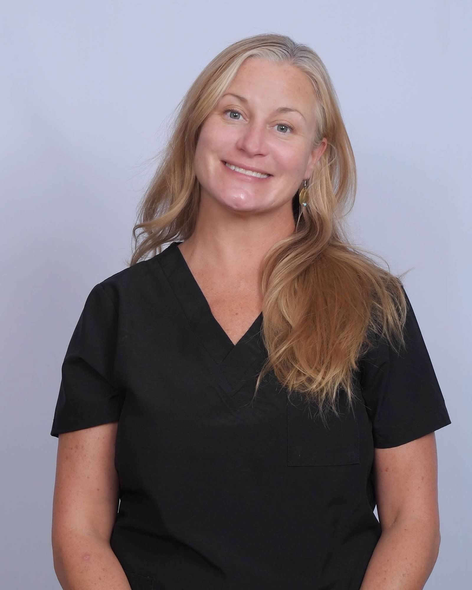 Woman with blonde hair wearing a black scrub top, smiling, against a light gray background.
