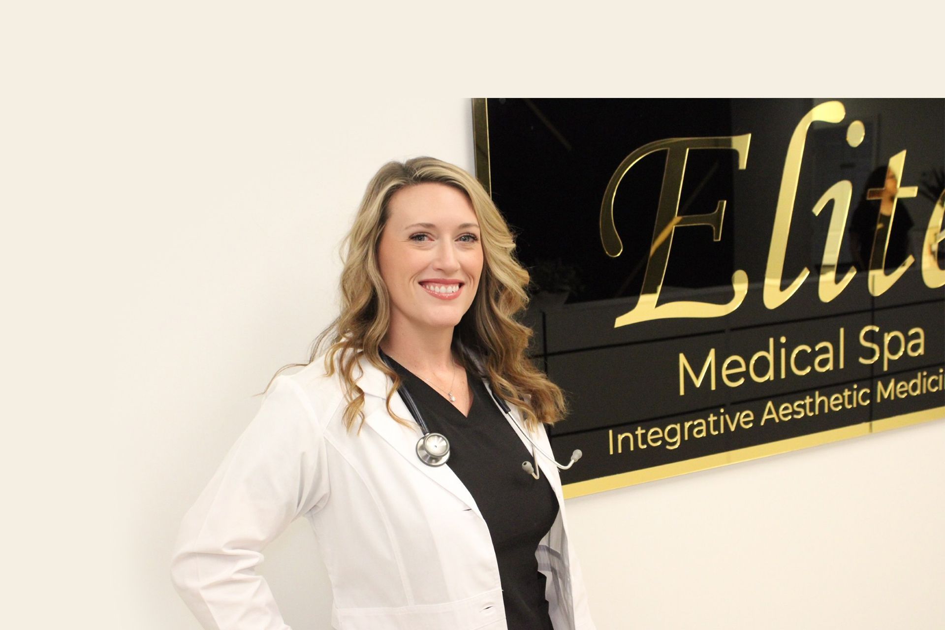 Woman in white coat smiles, stethoscope around neck, standing by 