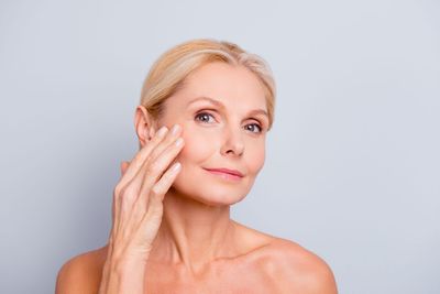 Woman gently applying skincare cream to her cheek against a light gray background