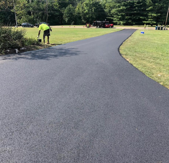 A worker in a bright yellow shirt stands on the edge of a newly paved, smooth black driveway in a grassy yard.