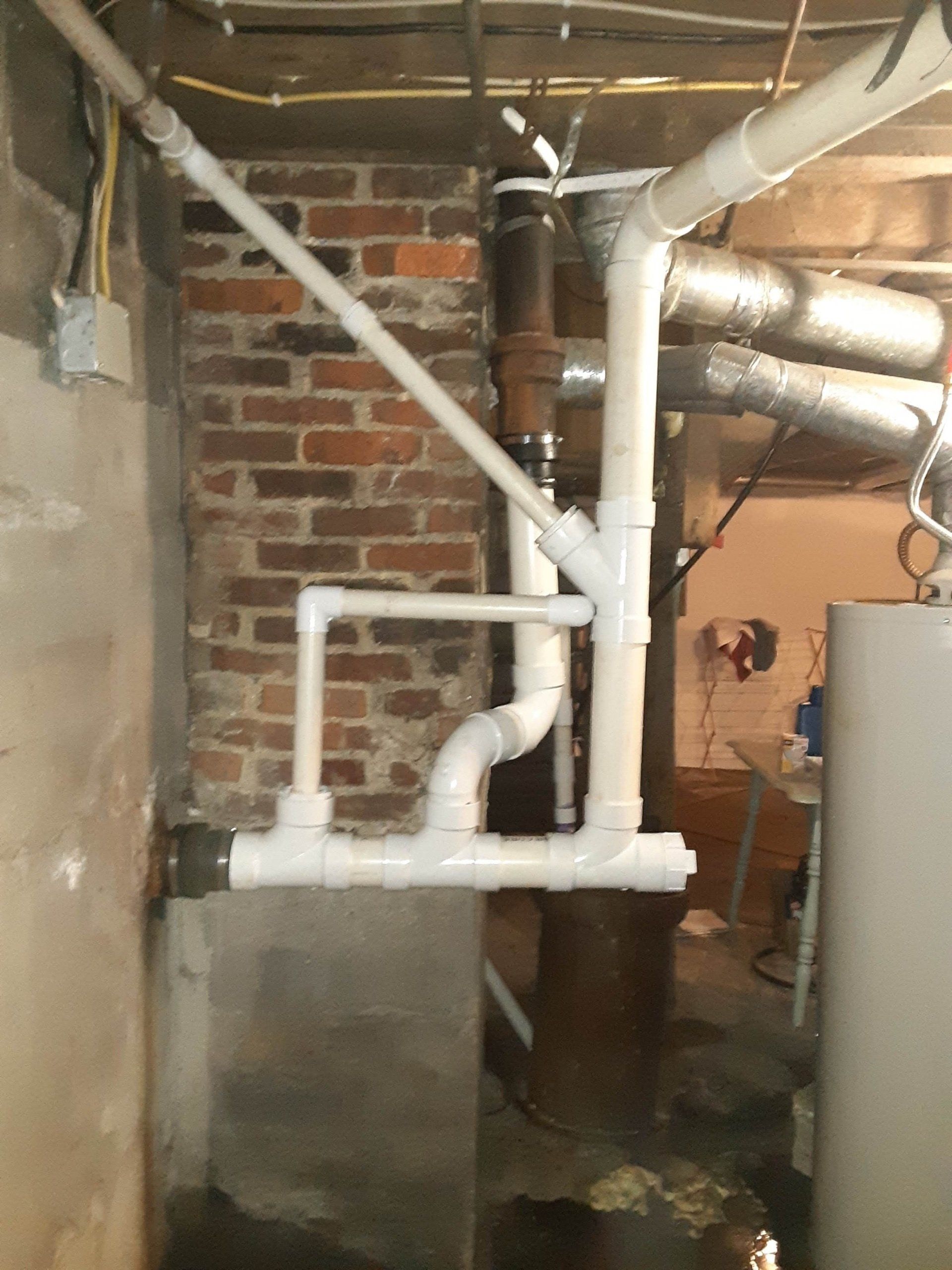 White PVC pipes and vents against a brick wall and concrete foundation in a basement.