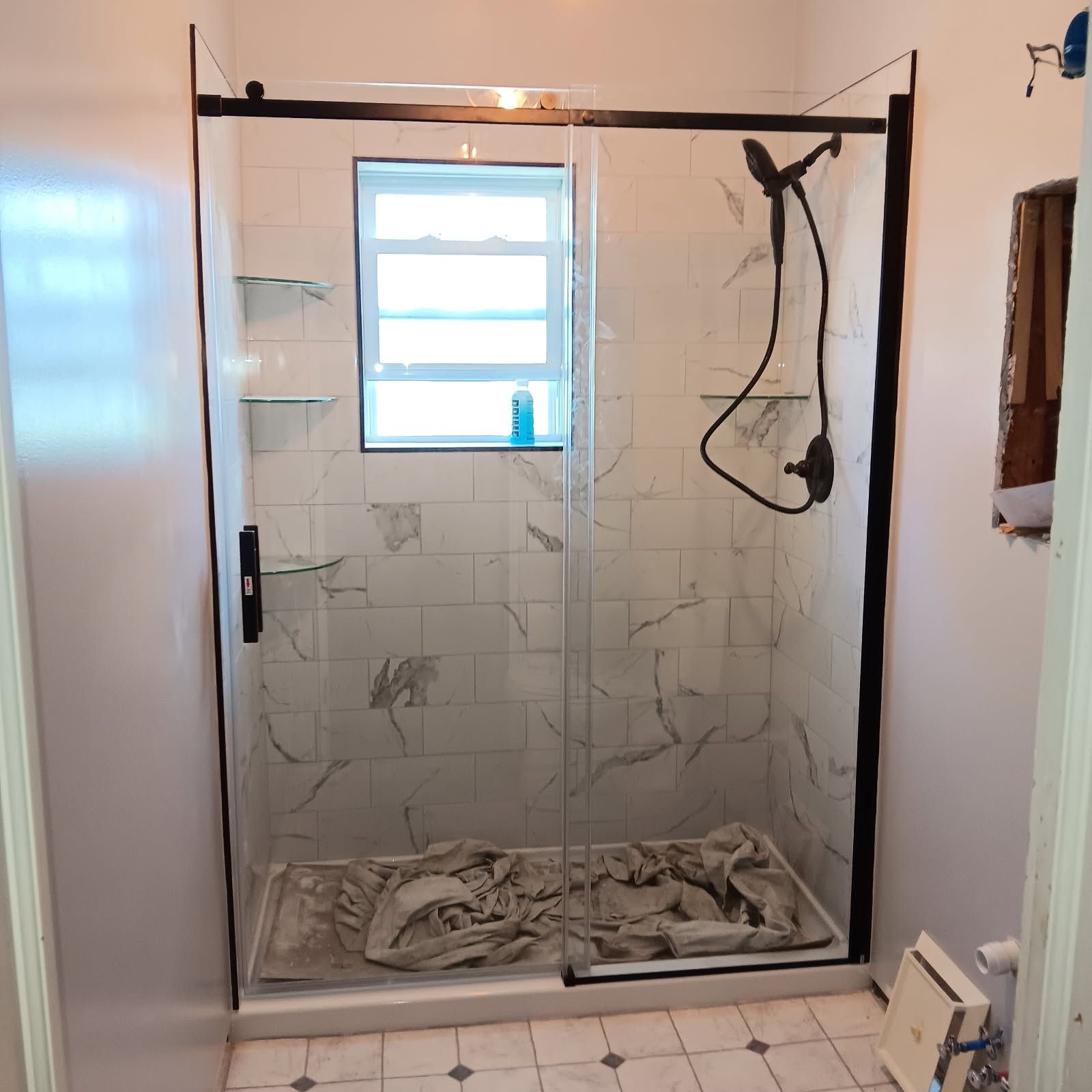 Black-framed glass shower in a bathroom with marble-like tiles and a small window; gray floor covering inside.