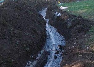 A narrow trench dug in soil, lined with gravel, likely for utility lines, outdoors