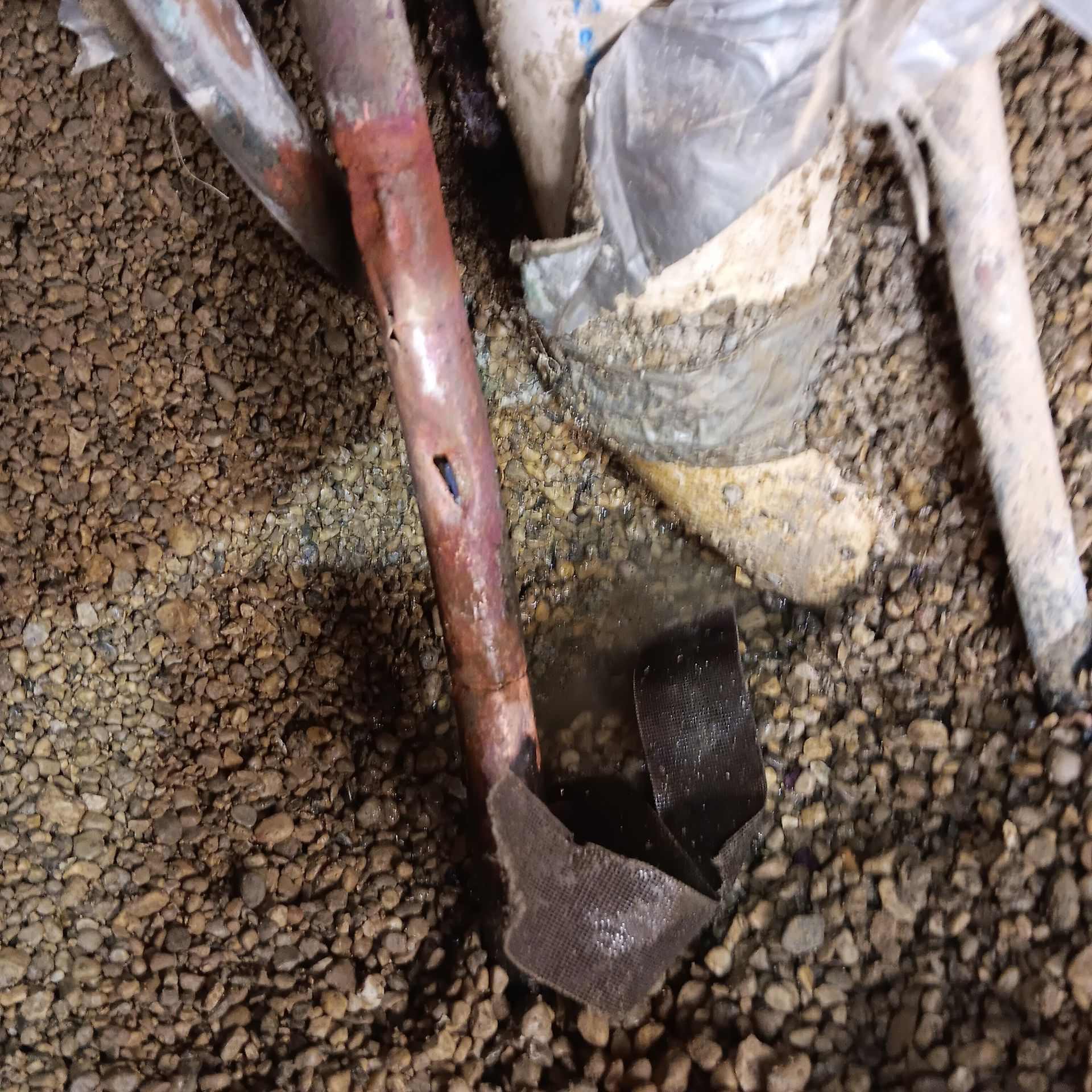 Damaged copper pipe with a hole, leaking water in a gravel-filled basement.