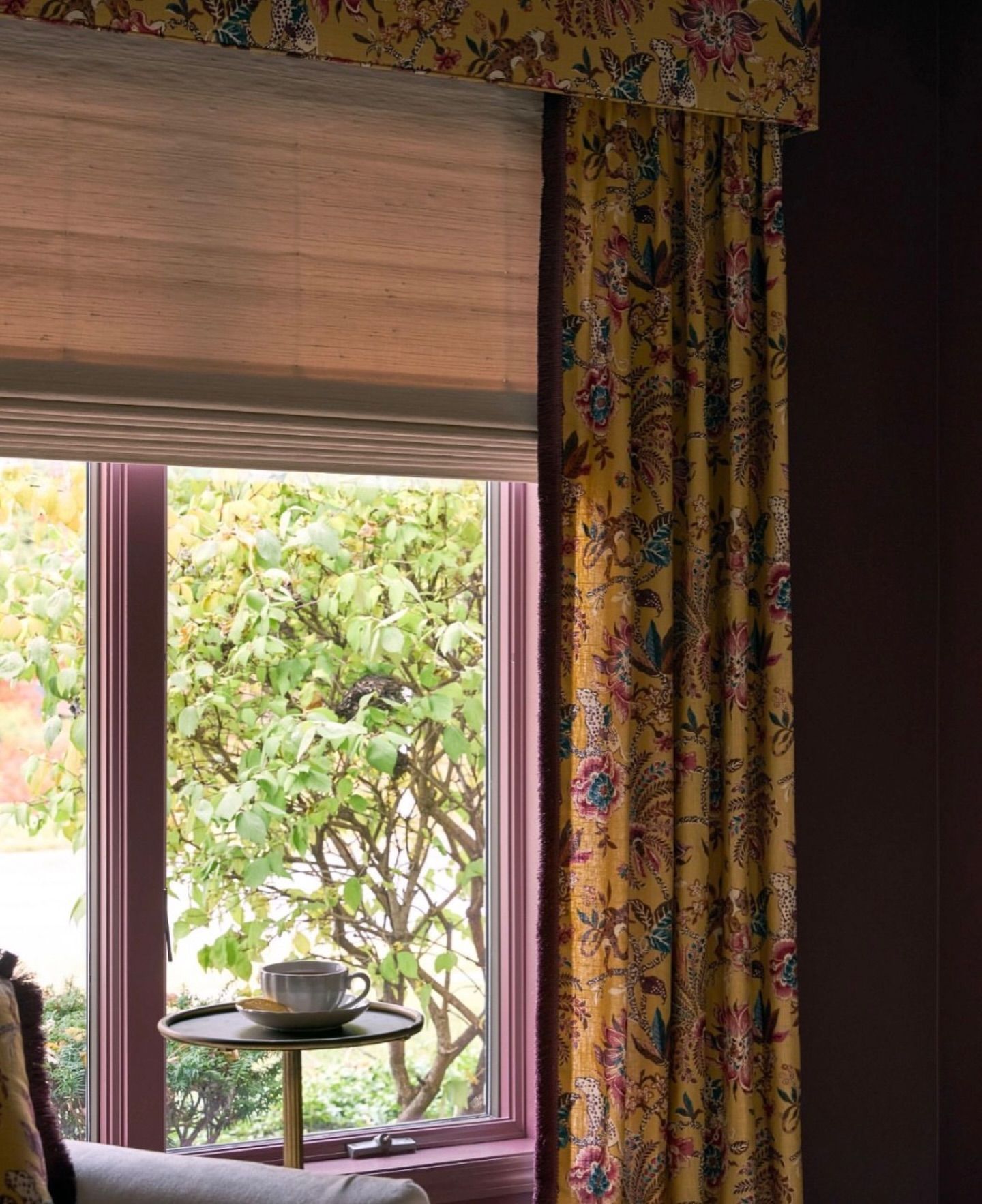 Window with mustard-yellow floral curtains and a matching valance, a tan Roman shade, and a small table with a teacup.