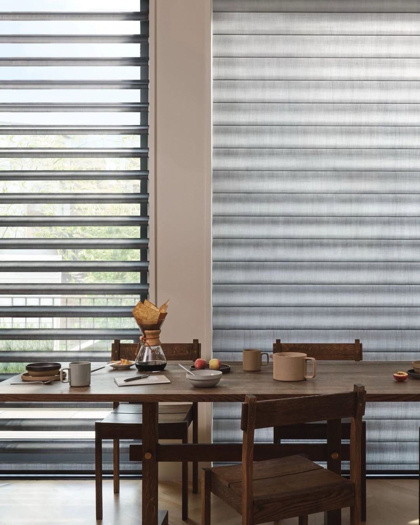 Wooden dining table with chairs in front of two windows covered by horizontal blinds. Coffee pot and mugs sit on the table.