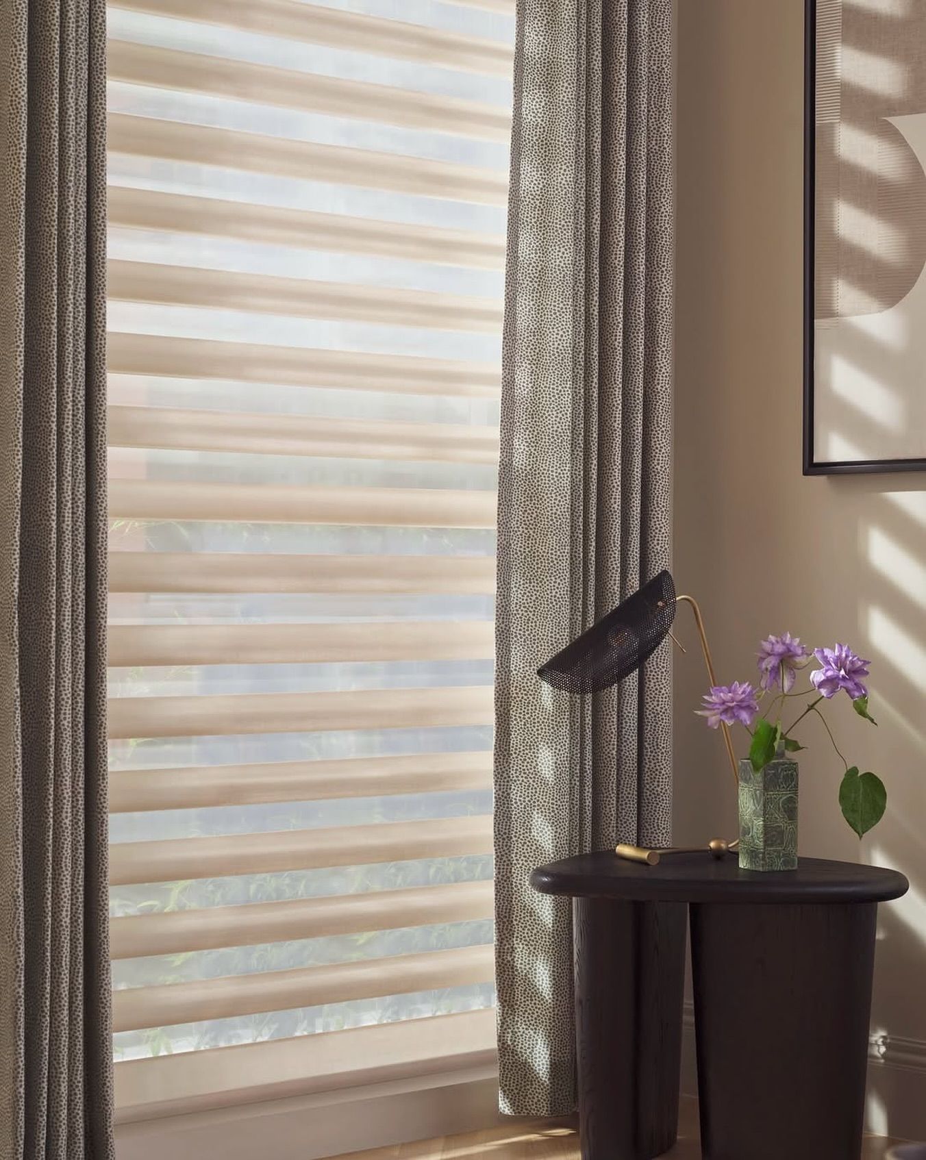 Window with layered beige blinds, patterned curtains, side table with flowers, and artwork.