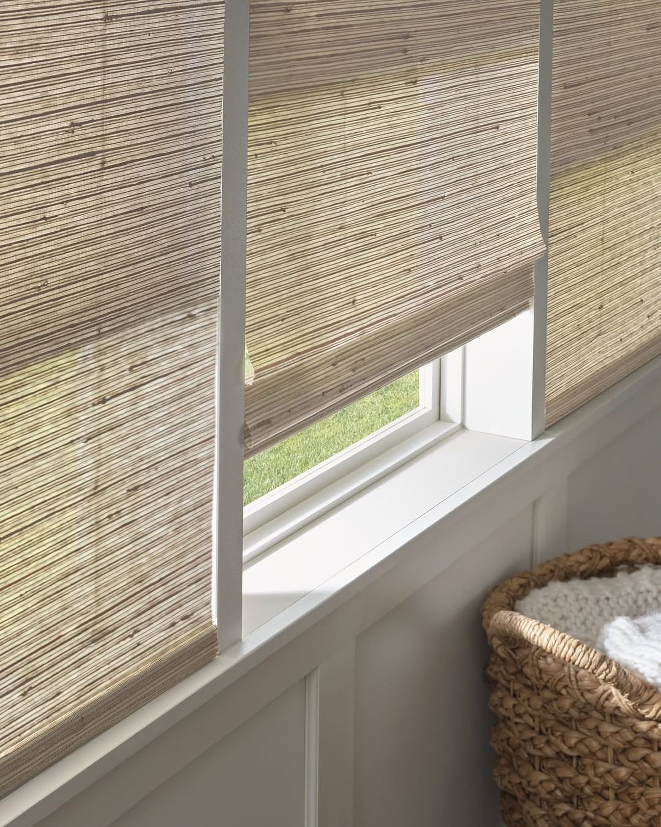 Woven window shades, partially lowered, over a white windowsill with a wicker basket in the foreground.