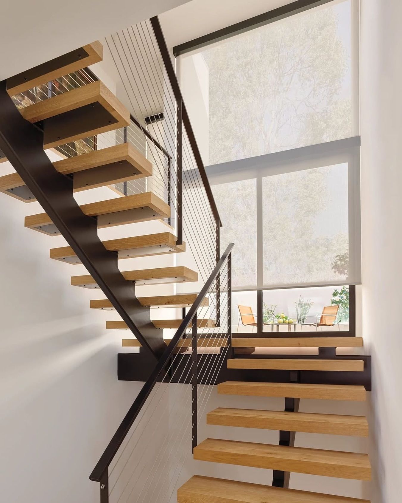 Modern wooden staircase with black metal supports and railing, near a window overlooking a patio.