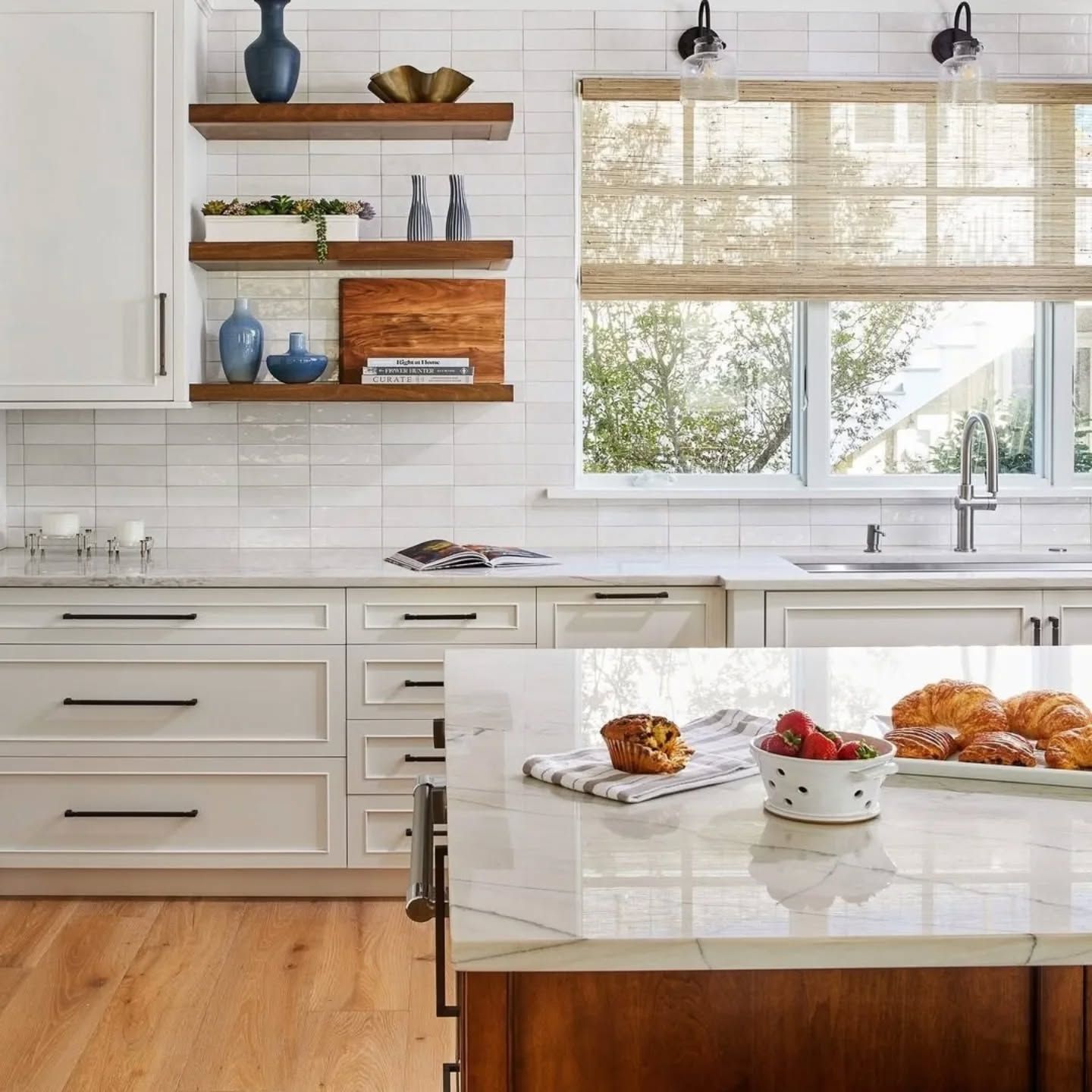 Bright kitchen with white cabinets, wood shelves, and a window with a woven shade. Island with breakfast.