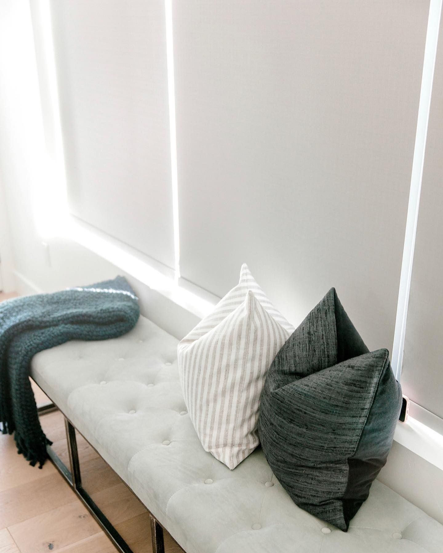 Gray bench with two pillows and a folded blanket beneath white window blinds.